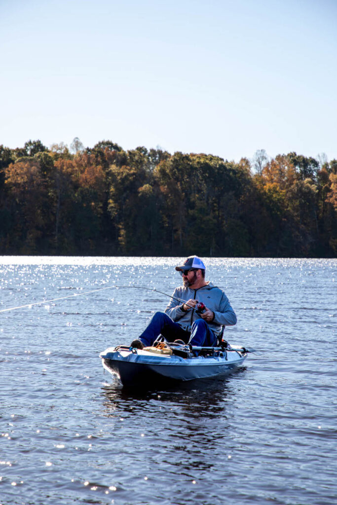 A man wearing a baseball cap and sunglasses fishing from a kayak on a large, sunlit lake, with a colorful autumn forest in the background.
