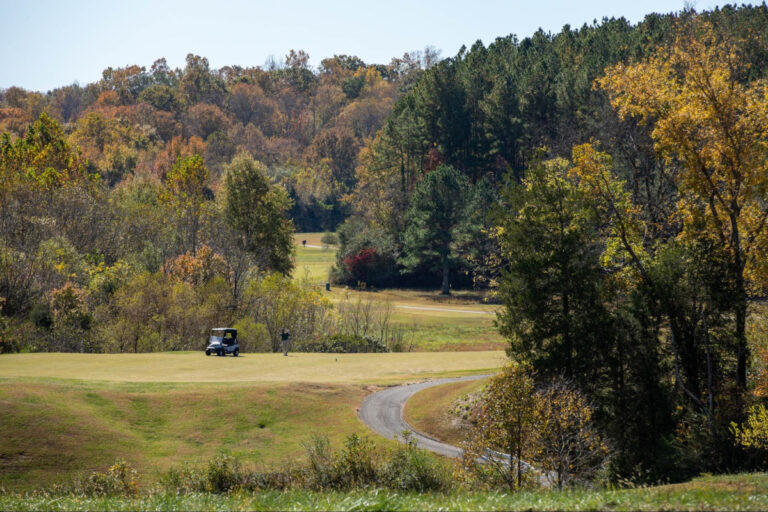 A golfer stands on a green tee box near a golf cart at the Manor Golf Club surrounded by colorful autumn trees.