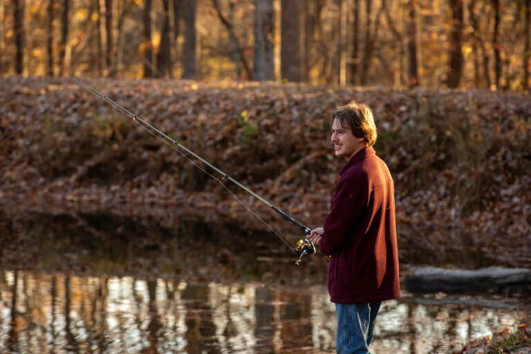 A young man wearing a maroon jacket and blue jeans stands near the edge of a body of water, smiling while holding a fishing rod in a wooded area with autumn trees in the background.